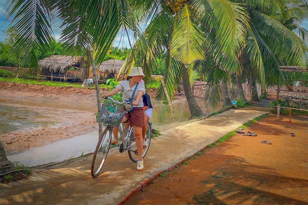 Tourist cycling under coconut palms on Con Chim Island, Tra Vinh, exploring village lanes and local life in the Mekong Delta