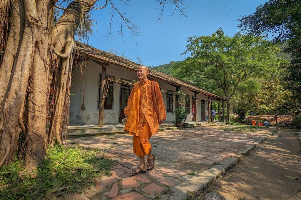 Theravada monk in orange robes walking past traditional wooden buildings at Huyen Khong Son Thuong Pagoda in Hue, Vietnam.