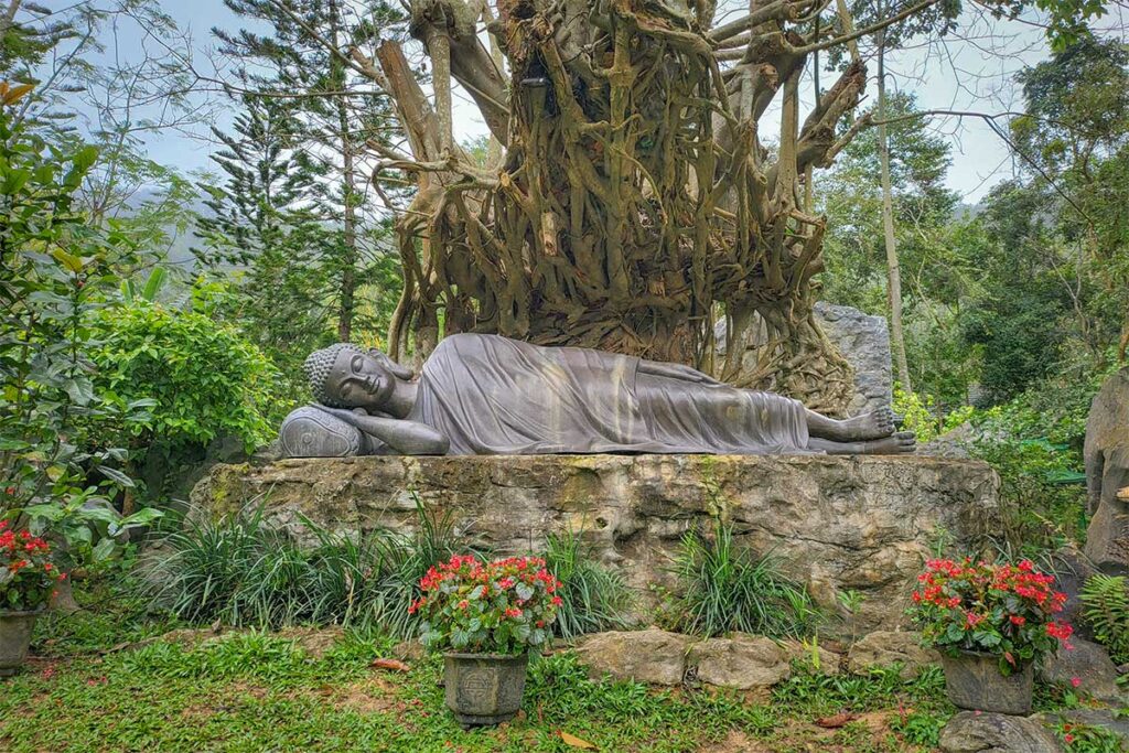 Reclining Buddha statue beneath a banyan tree in the gardens of Huyen Khong Son Thuong Pagoda, symbolizing peace and enlightenment in Hue’s countryside.