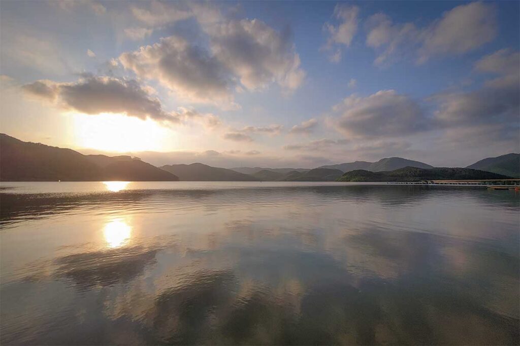 Golden sunset reflecting over Khe Ngang Lake near Hue, with calm waters and mountain silhouettes creating a peaceful countryside scene.