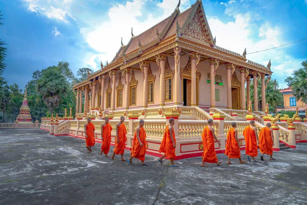 Monks in orange robes walking past the main hall of Kh’leang Pagoda in Soc Trang, a 500-year-old Khmer Buddhist temple with ornate golden details.