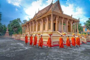 Monks in orange robes walking past the main hall of Kh’leang Pagoda in Soc Trang, a 500-year-old Khmer Buddhist temple with ornate golden details.