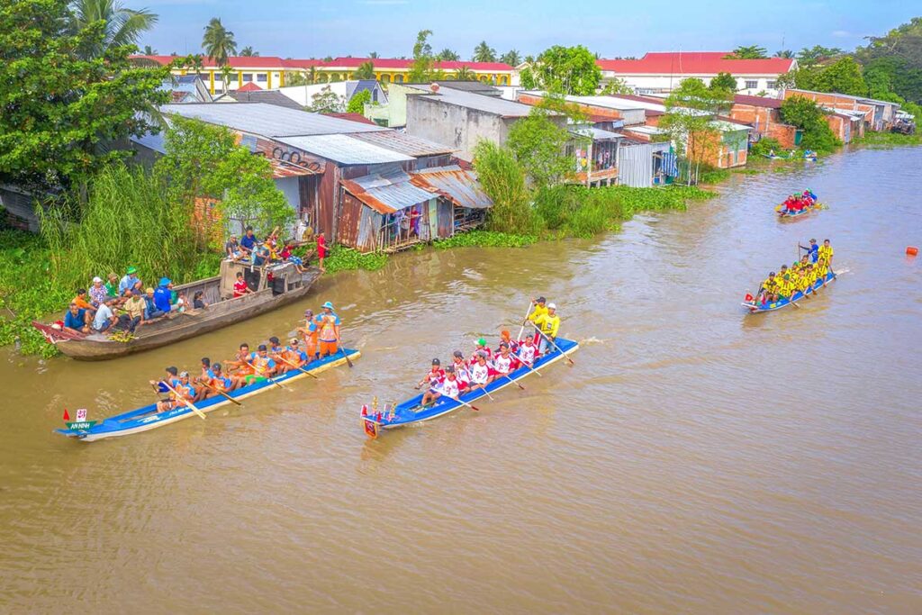 Khmer rowing teams racing along a rural canal during the Ghe Ngo Boat Race Festival in Soc Trang, passing local houses and boats filled with spectators.