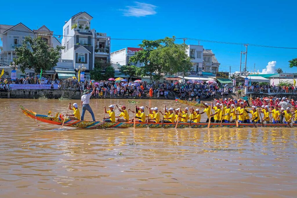 Rowers in yellow uniforms competing in the Ghe Ngo Boat Race Festival on the Maspero River, Soc Trang, with large crowds watching from the riverside.