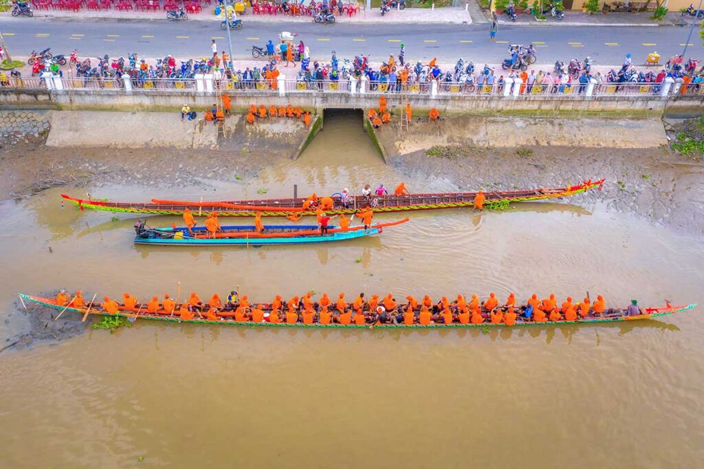 Khmer pagoda teams in orange robes preparing traditional longboats before the Ghe Ngo Boat Race Festival in Soc Trang, viewed from above along the riverbank.