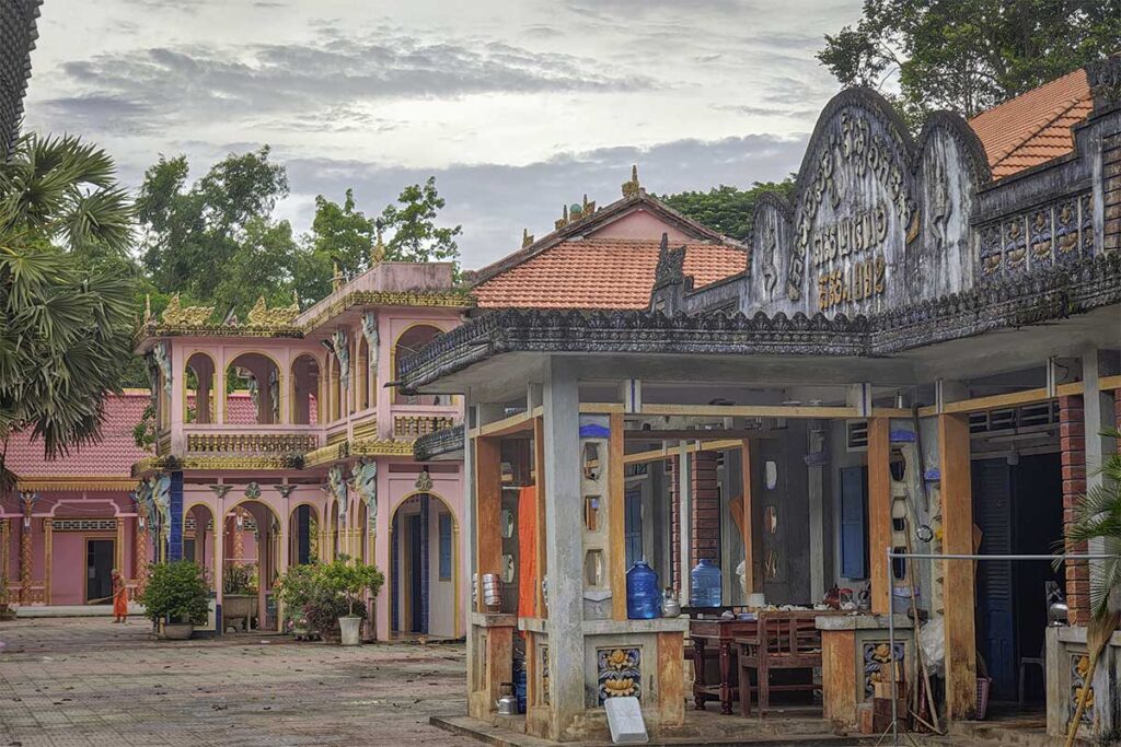 Courtyard of Nodol Pagoda in Tra Vinh with Khmer-style buildings and a monk walking past pink and gold decorated halls