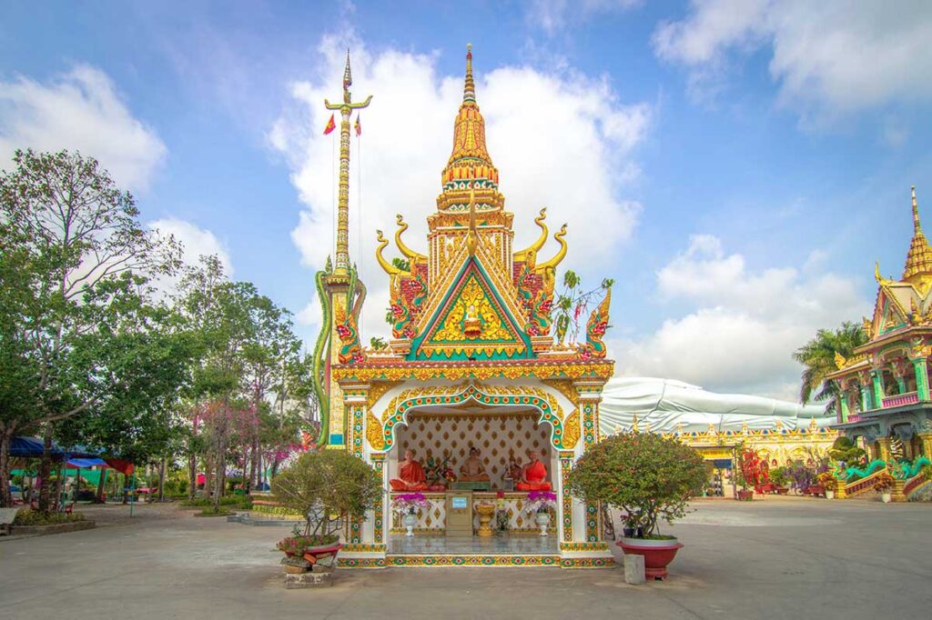 Small pavilion shrine at Som Rong Pagoda with seated Buddha statues, colorful Khmer roof details, and the reclining Buddha visible in the background