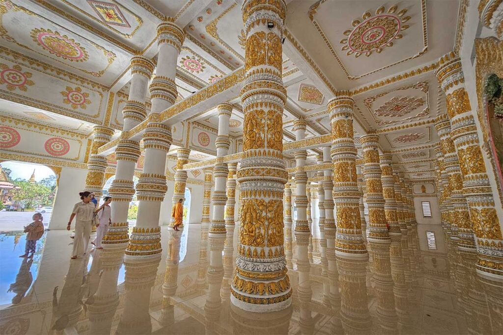 Interior beneath the reclining Buddha statue at Som Rong Pagoda, with white walls and ornate golden columns on a polished floor