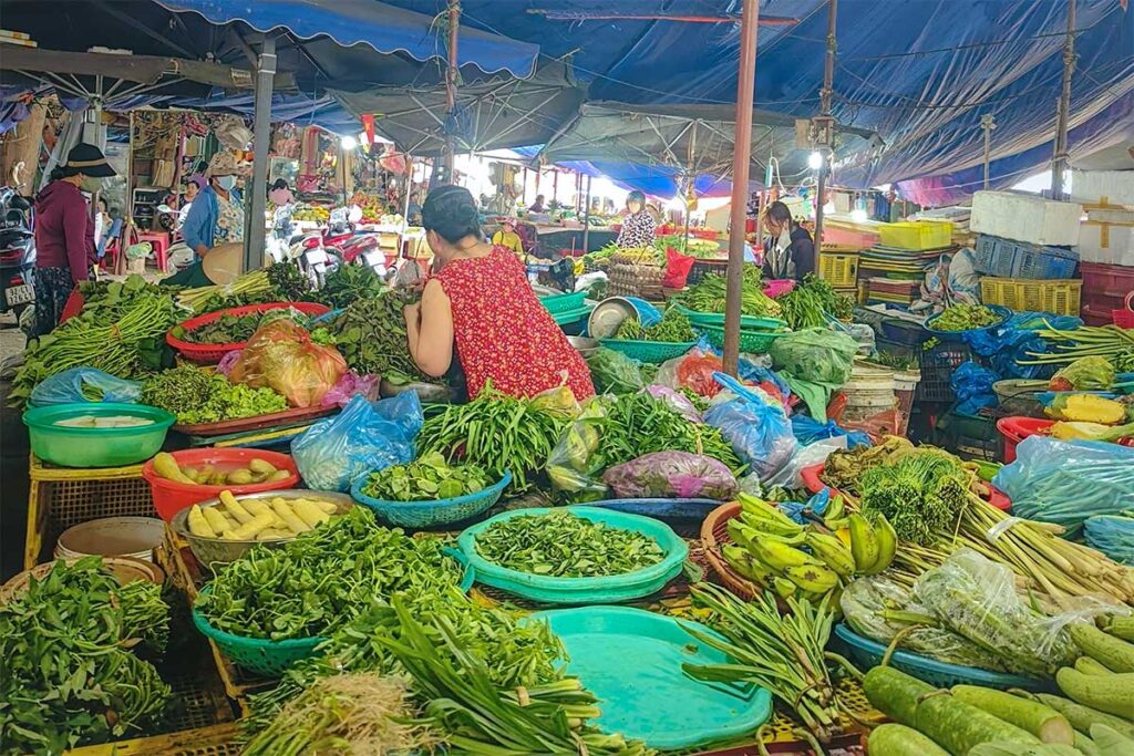 Fresh vegetables at Tan An Market Hoi An with baskets of herbs, greens, and bananas under tarpaulin stalls.