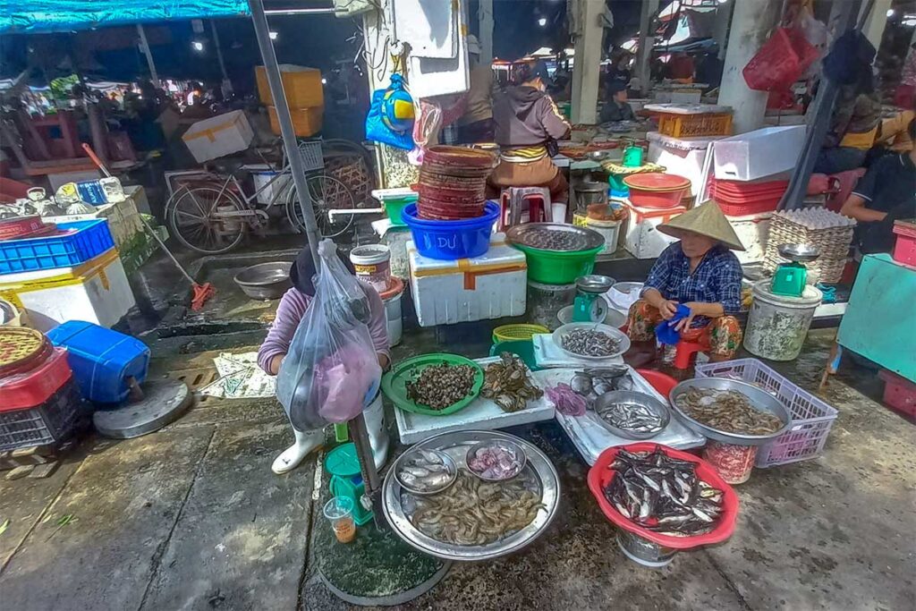 Seafood section of Tan An Market Hoi An with women selling shrimp, fish, and shellfish in metal trays