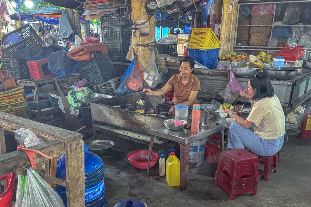 Local vendor cooking noodles at a small food stall inside Tan An Market Hoi An with customers eating on low stools.