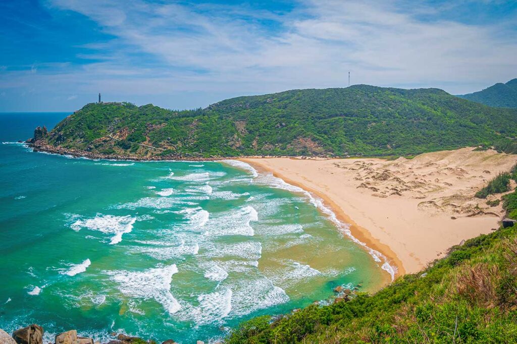 View of Bai Mon Beach in Phu Yen, with golden sand, blue waves, and Mui Dien Lighthouse on the hilltop.