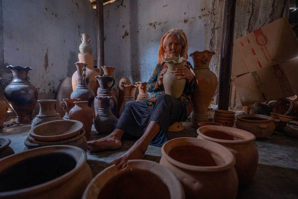 Elderly Cham woman sitting among handmade terracotta pots, inspecting her work inside Bau Truc Pottery Village.