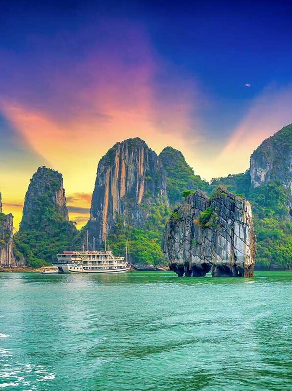 Halong Bay tours – Local Vietnam | cruise boat sailing between limestone karsts at sunset on Vietnam’s UNESCO World Heritage bay