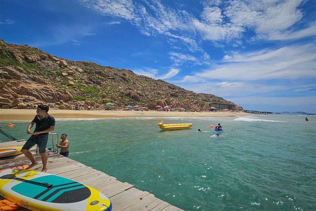 Water sports at Nhon Hai – Locals and travelers enjoying paddleboards and a banana boat on the calm sea framed by rocky hillsides.