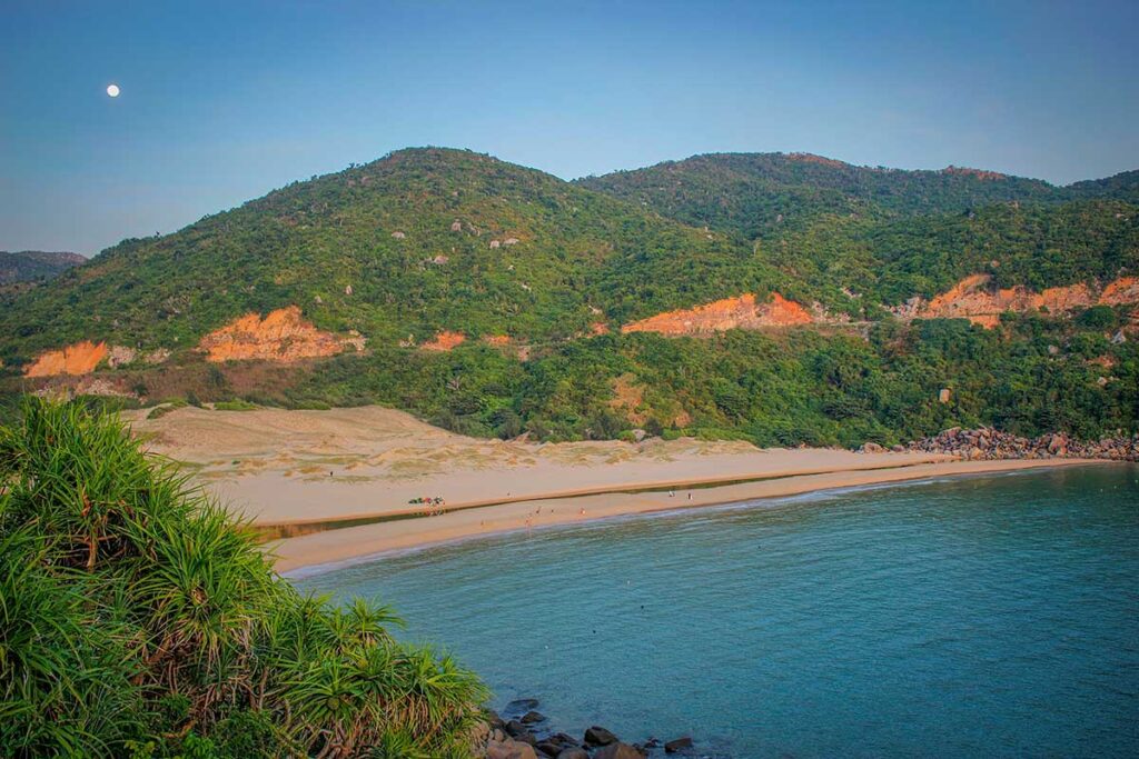 View of Bai Mon Beach from Mui Dien Lighthouse, showing sandy dunes, green hills, and calm blue water.