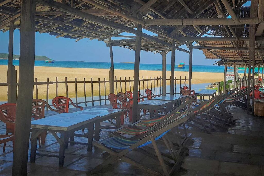 Seaside restaurant at Long Thuy Beach – Simple beachfront eatery with plastic chairs and hammocks overlooking the sandy shore and blue sea.