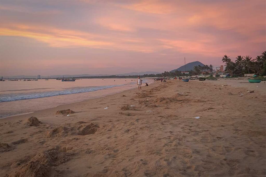 Evening atmosphere on Long Thuy Beach – Locals and visitors walking along the sand at sunset, with pastel skies and fishing boats offshore.