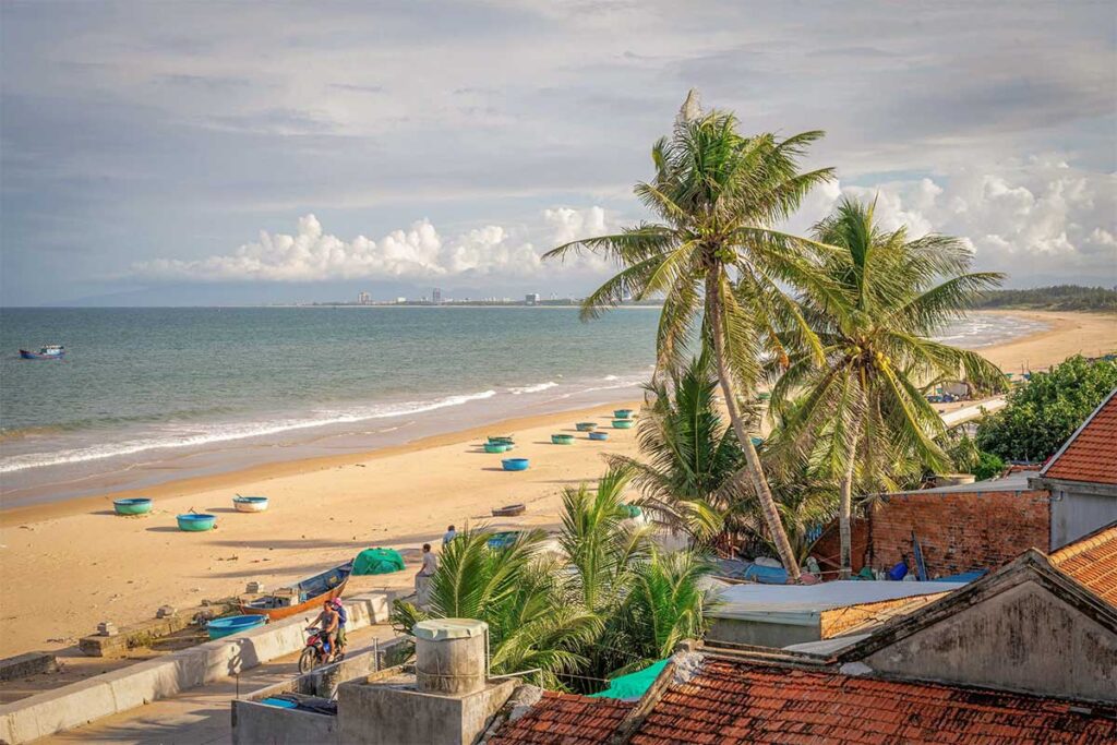 Long Thuy Beach coastline with fishing coracles – Wide sandy beach lined with round fishing boats and coconut palms, looking out toward Tuy Hoa city on the horizon.