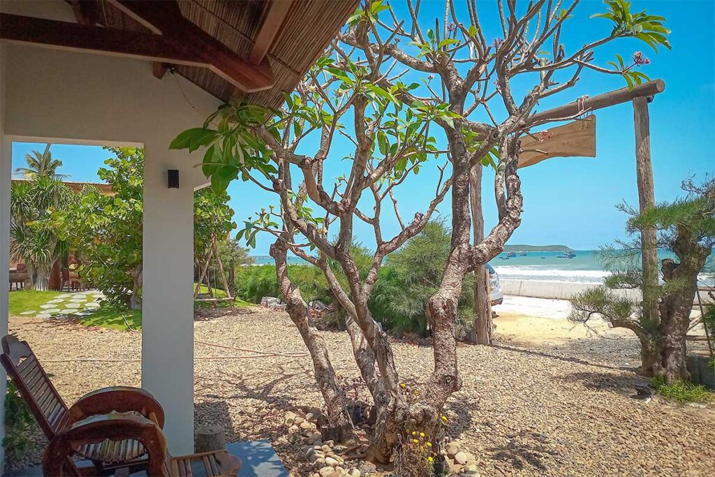 Seaside homestay at Long Thuy Beach with garden trees and a shaded porch, overlooking the sandy beach and fishing boats on the Phu Yen coast.