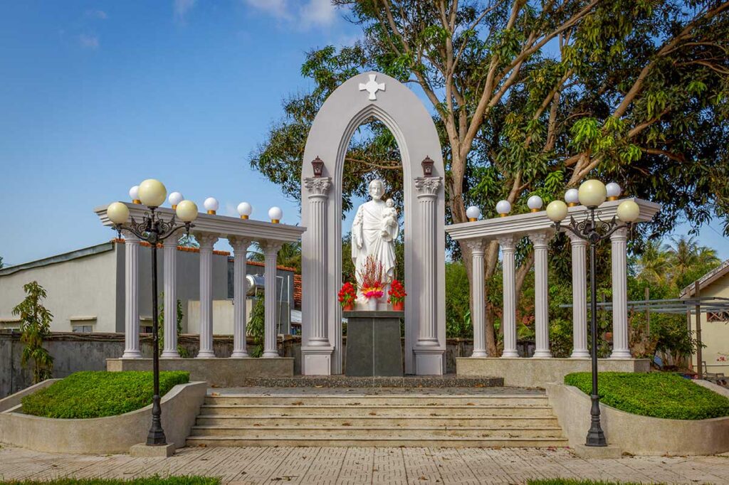 Marble statue of Jesus in the courtyard of Mang Lang Church, framed by classical columns and archways under blue skies.