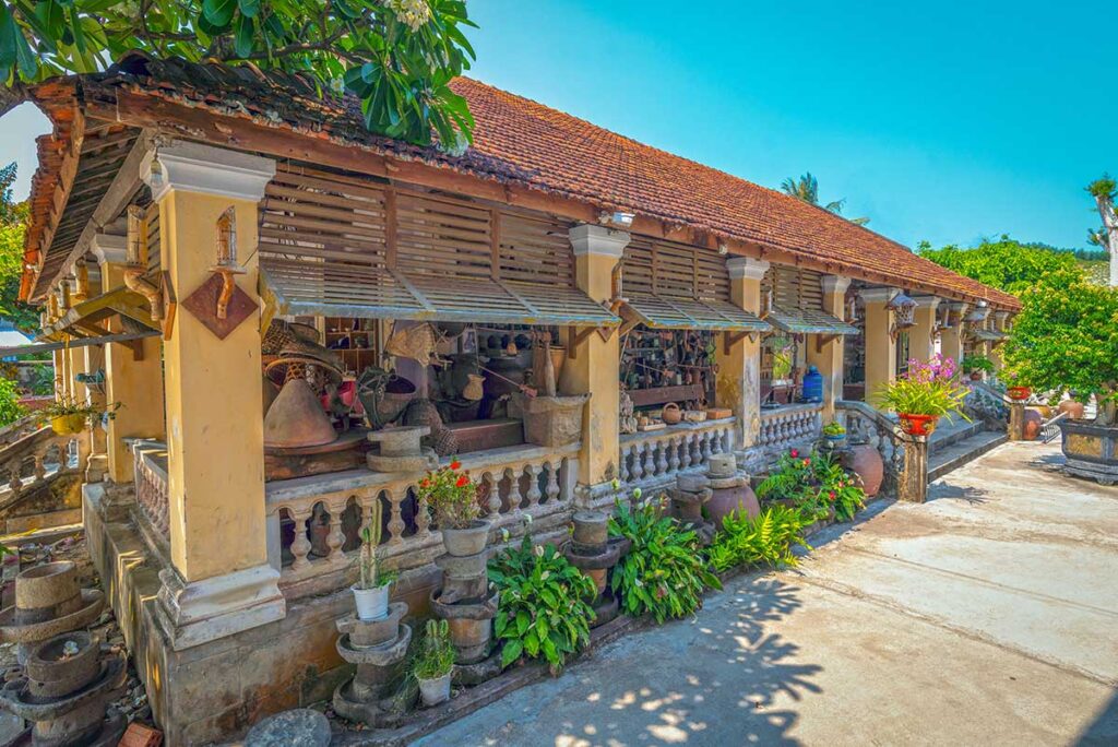 Old house beside Mang Lang Church displaying antiques, pots, and quirky memorabilia under a tiled roof.