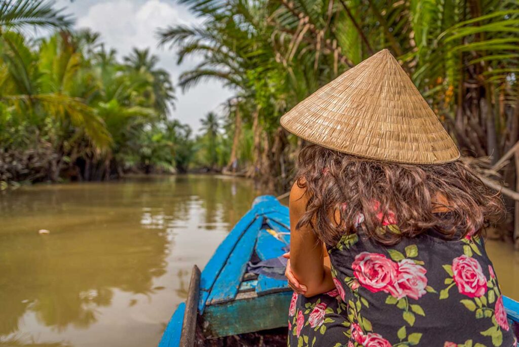 Mekong Delta tours – Local Vietnam | traveler wearing a conical hat on a traditional boat through palm-lined canals in southern Vietnam