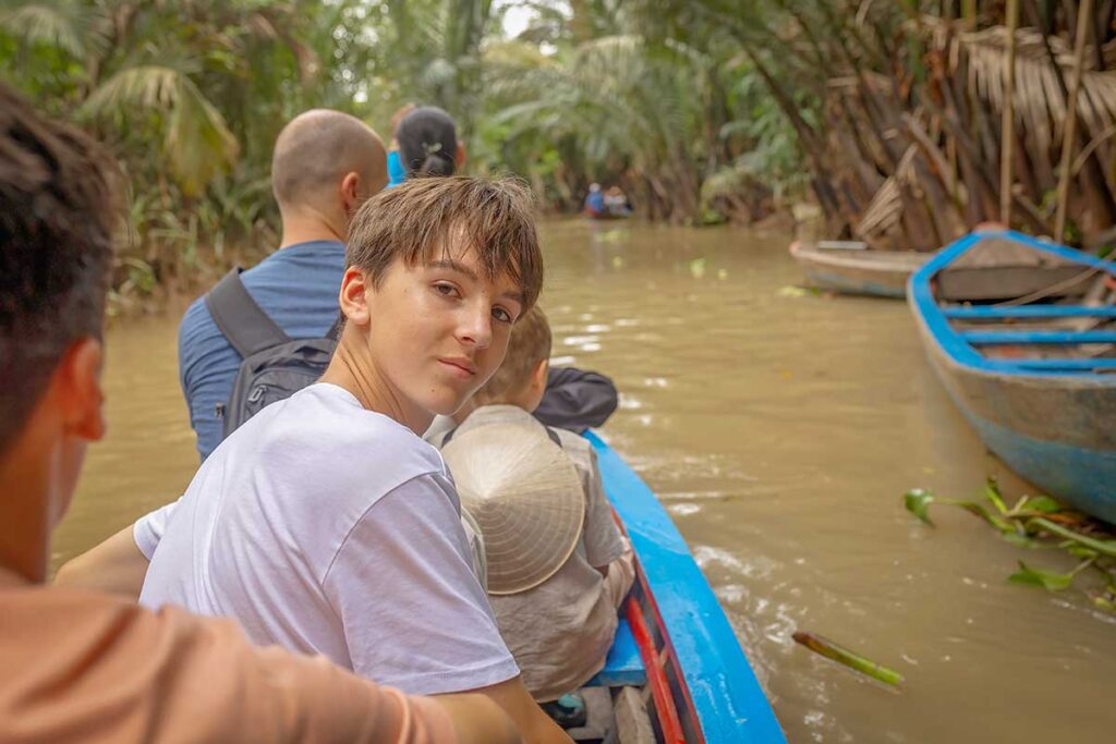 Teenager sitting in a sampan navigating narrow canals of the Mekong Delta, a memorable adventure for families with older kids.