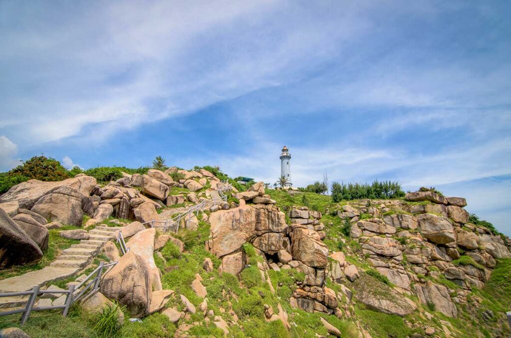 Stone staircase leading up to Mui Dien (Dai Lanh) Lighthouse surrounded by rugged rocks and green slopes.