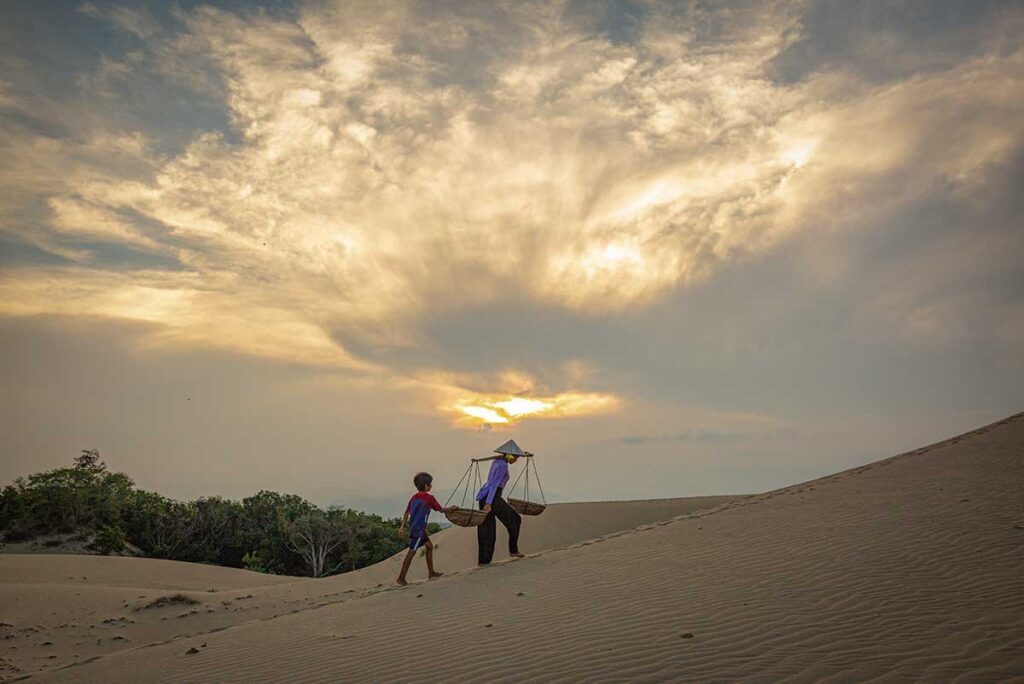 Local woman with conical hat and child walking over Nam Cuong Sand Dunes at dusk