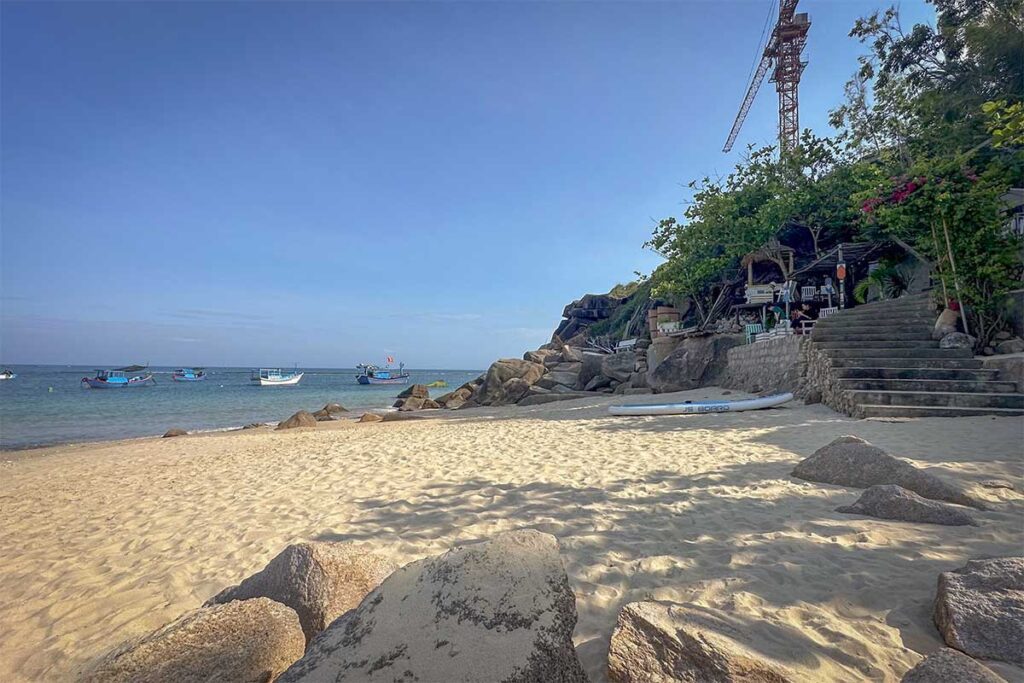 Quiet stretch of Nhon Hai Beach – Golden sand, scattered rocks, and fishing boats anchored offshore near a rustic beach café.