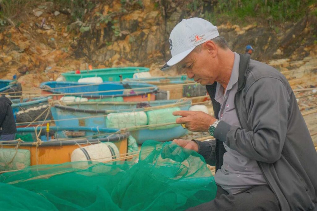 Fisherman repairing green nets beside colorful round basket boats in Nhon Ly fishing village, Quy Nhon.