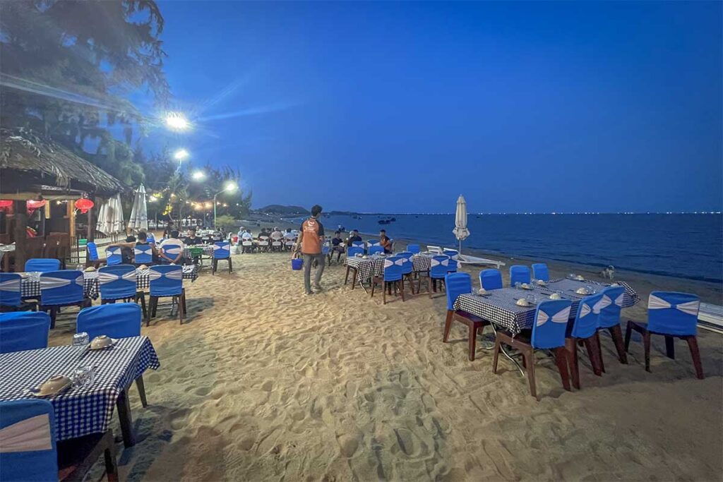 Open-air seafood restaurant set directly on the sand at Ninh Chu Beach, Phan Rang, with tables overlooking the sea at night.