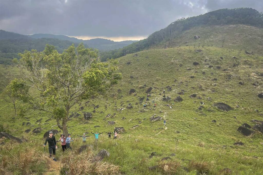 Group of trekkers hiking a grassy valley trail in Nui Chua National Park, passing scattered rocks under cloudy skies.