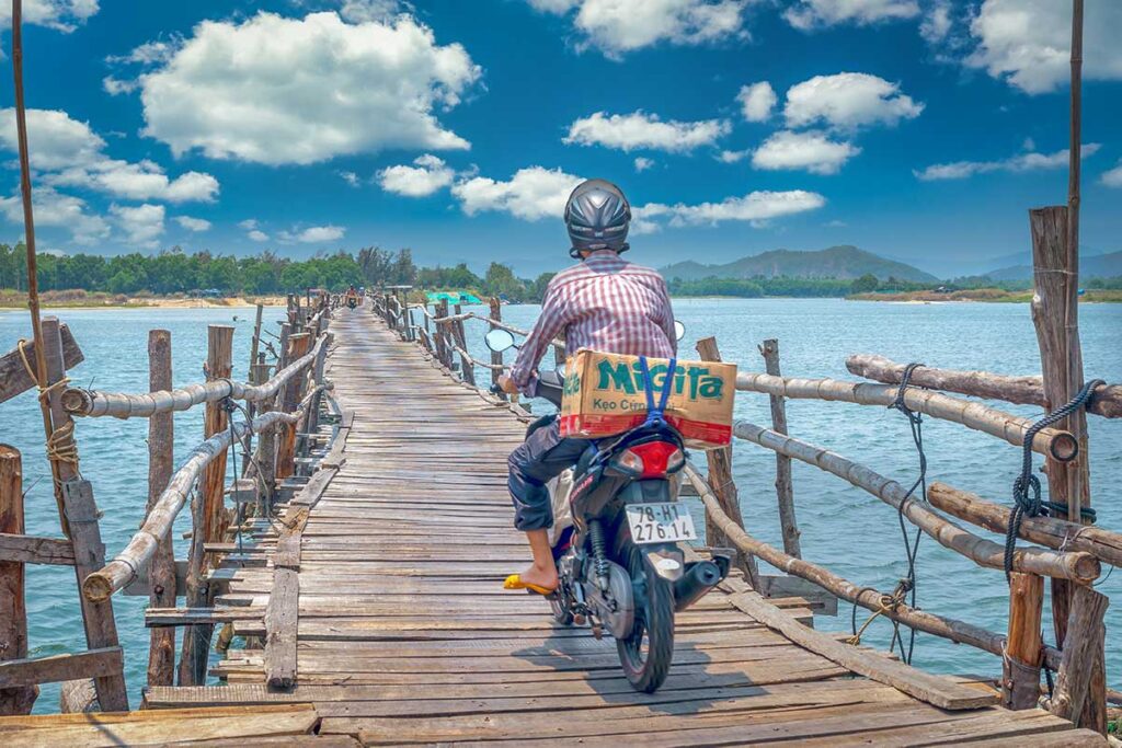 Local man riding a motorbike across Ong Cop Wooden Bridge in Phu Yen, Vietnam, with bright blue skies and clouds above the river.
