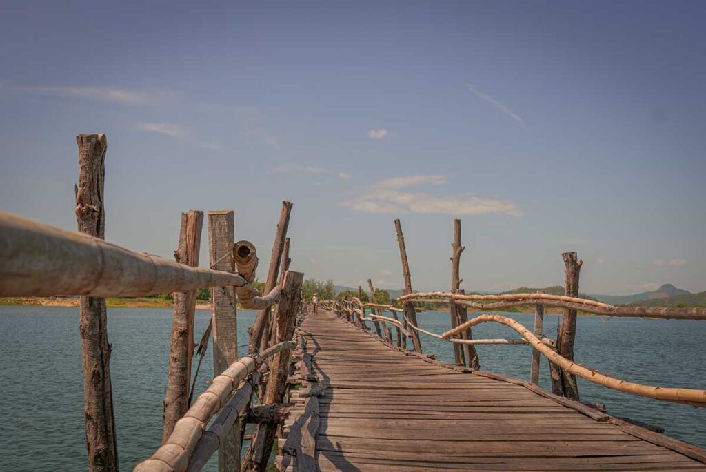 Perspective along Ong Cop Wooden Bridge in Phu Yen, Vietnam, showing the bamboo rails and rustic wooden planks crossing the river.