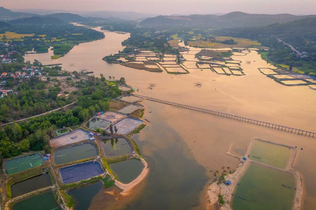 Drone photo of Ong Cop Wooden Bridge stretching across the Binh Ba River estuary in Phu Yen, surrounded by aquaculture ponds and coastal scenery.