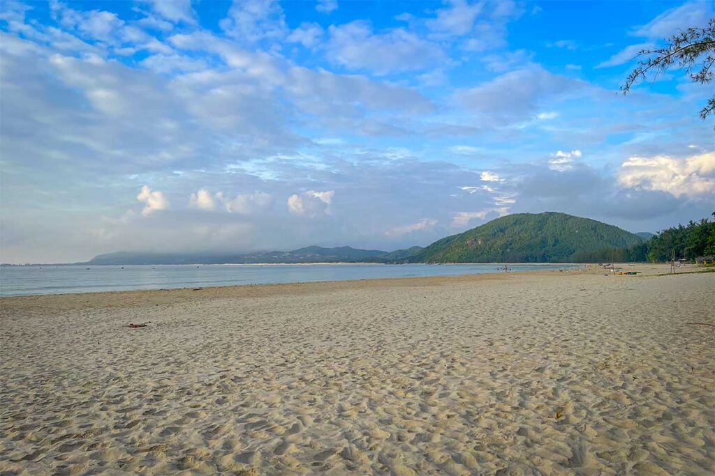 Wide, quiet sands at Tu Nham Beach with forested hills and gentle, swimmable water.