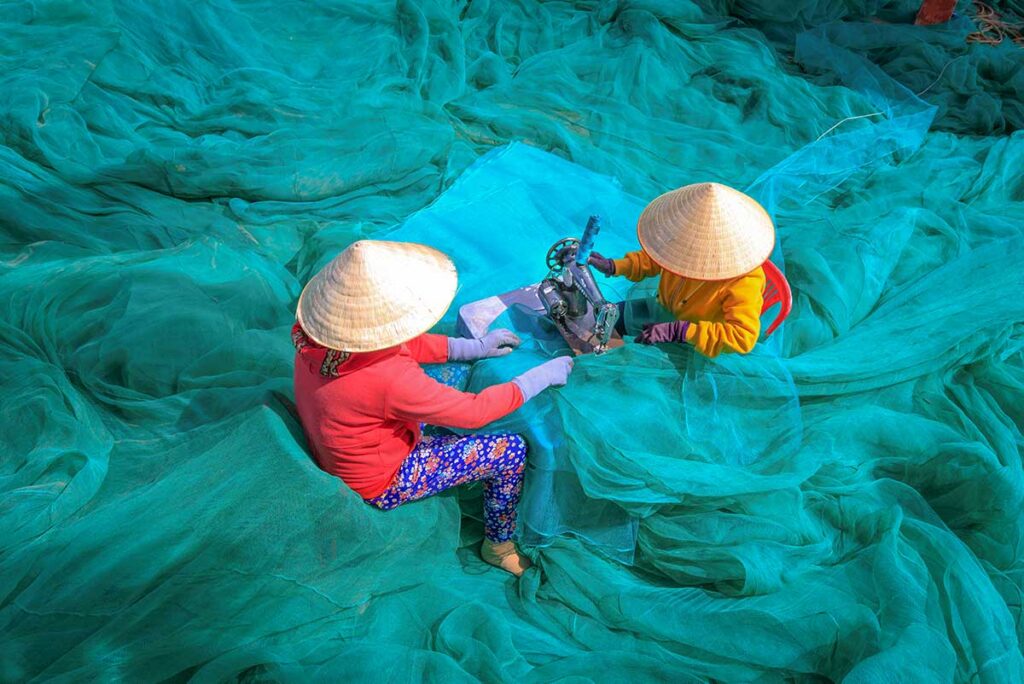 Women repairing fishing nets in Vinh Hy village – Two women in conical hats sewing green fishing nets by hand, a glimpse of daily life and traditional work in Vinh Hy fishing community.