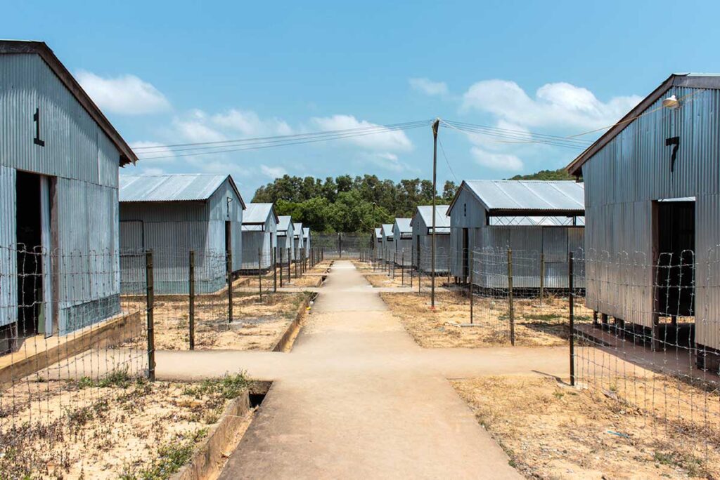 Row of numbered corrugated iron detention barracks stretching along a central pathway at Phu Quoc Prison, separated by barbed wire fencing on both sides.
