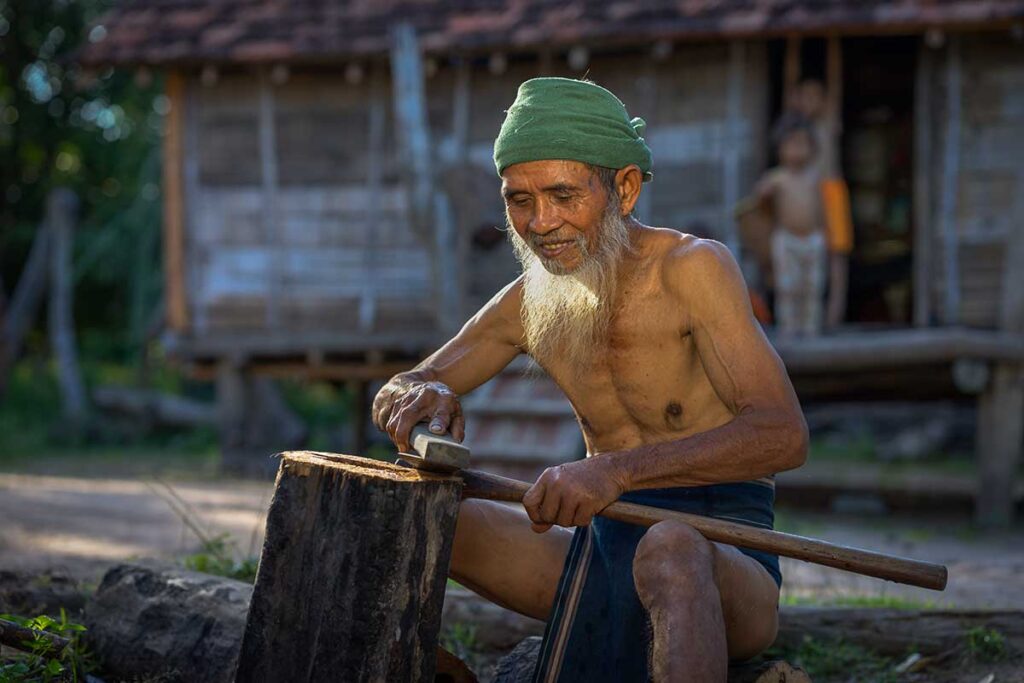 Local villager crafting wood outside a traditional stilt house in the Central Highlands, an authentic experience during rural Central Vietnam tours.