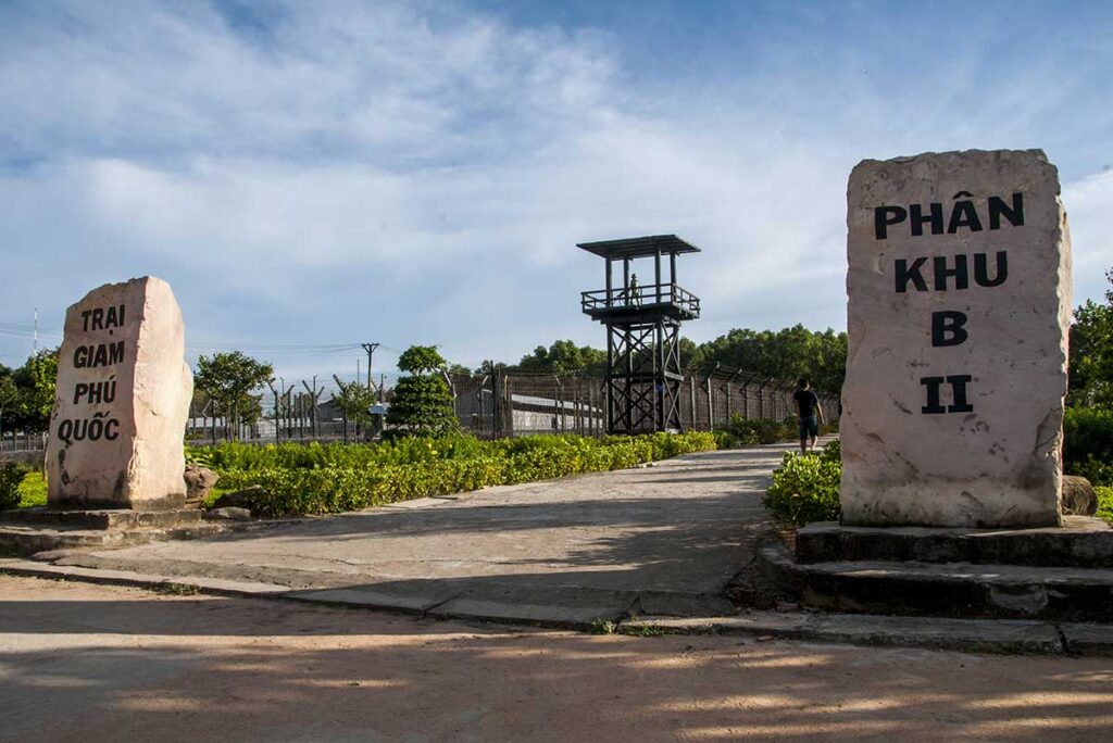 Entrance to Zone B2 of Phu Quoc Prison, marked by two large stone pillars and a watchtower silhouetted against the sky, with barbed wire fencing stretching along the perimeter.