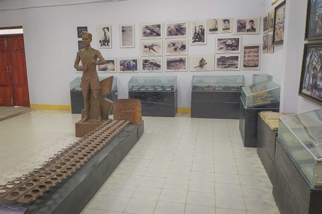 Interior of the exhibition house at Phu Quoc Prison showing display cases with wartime artifacts, a prisoner sculpture, a metal grating used for torture, and historical photographs covering the walls.