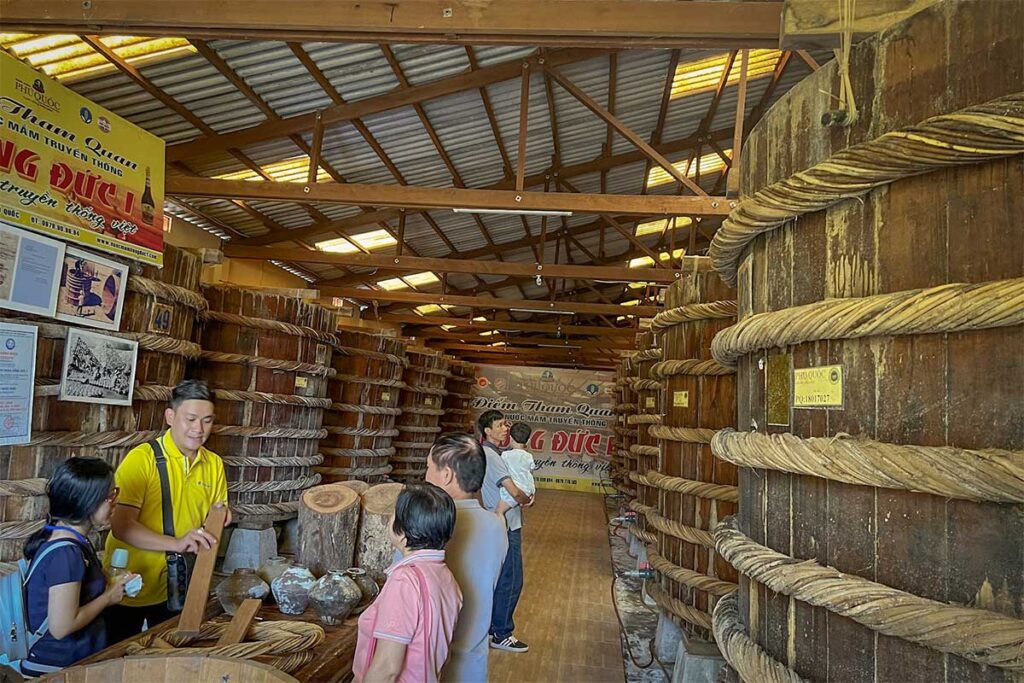 Visitors on a tour inside Hong Duc fish sauce factory in Phu Quoc, walking between rows of large traditional wooden fermentation barrels