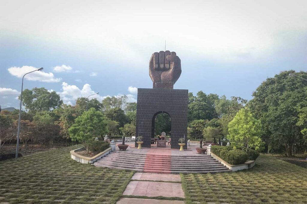 Large fist-shaped monument rising above a dark stone base at the prisoner graveyard near Phu Quoc Prison, surrounded by a well-kept garden and steps leading to an altar area.