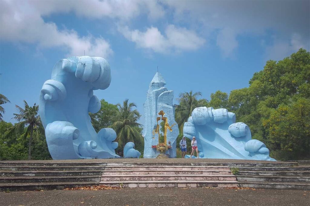 The Doi Sim martyrs memorial at Phu Quoc Prison, featuring large blue wave-shaped sculptures on either side of a central pointed pillar with a golden figure, set on a stepped platform surrounded by tropical trees.