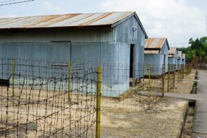 Rusted corrugated iron detention barracks at Phu Quoc Prison viewed from behind a barbed wire fence, showing the harsh and decayed conditions of the former Vietnam War prison camp.