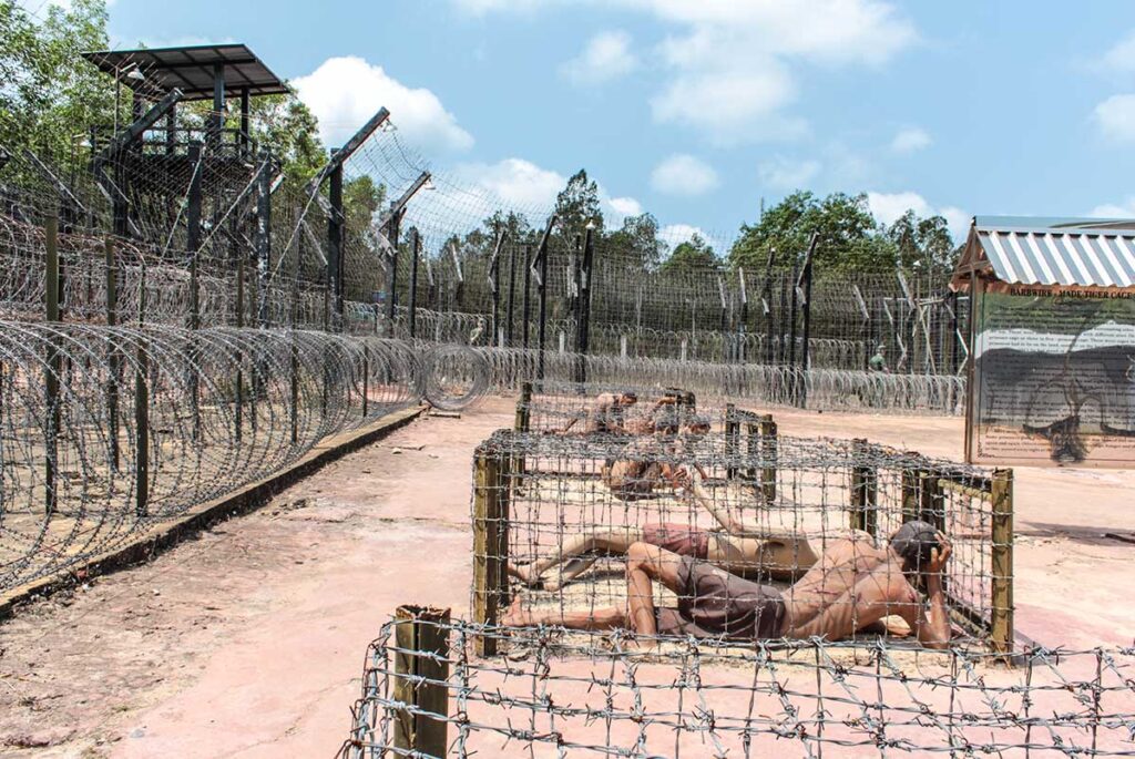 Wax figures of prisoners confined in barbed wire tiger cages at Phu Quoc Prison, with a watchtower and perimeter fencing visible in the background.