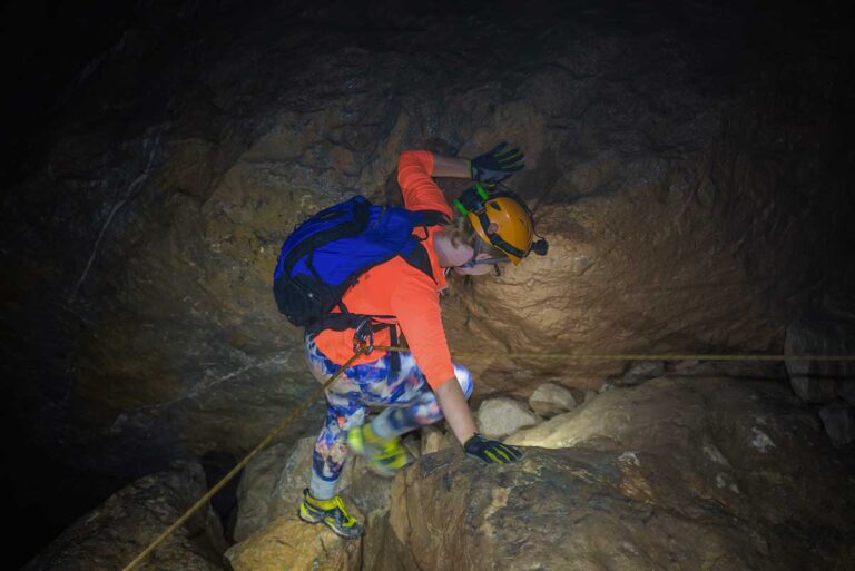 Best adventure activities in Vietnam, showing a traveler climbing inside a dark limestone cave in Phong Nha with helmet and safety rope.