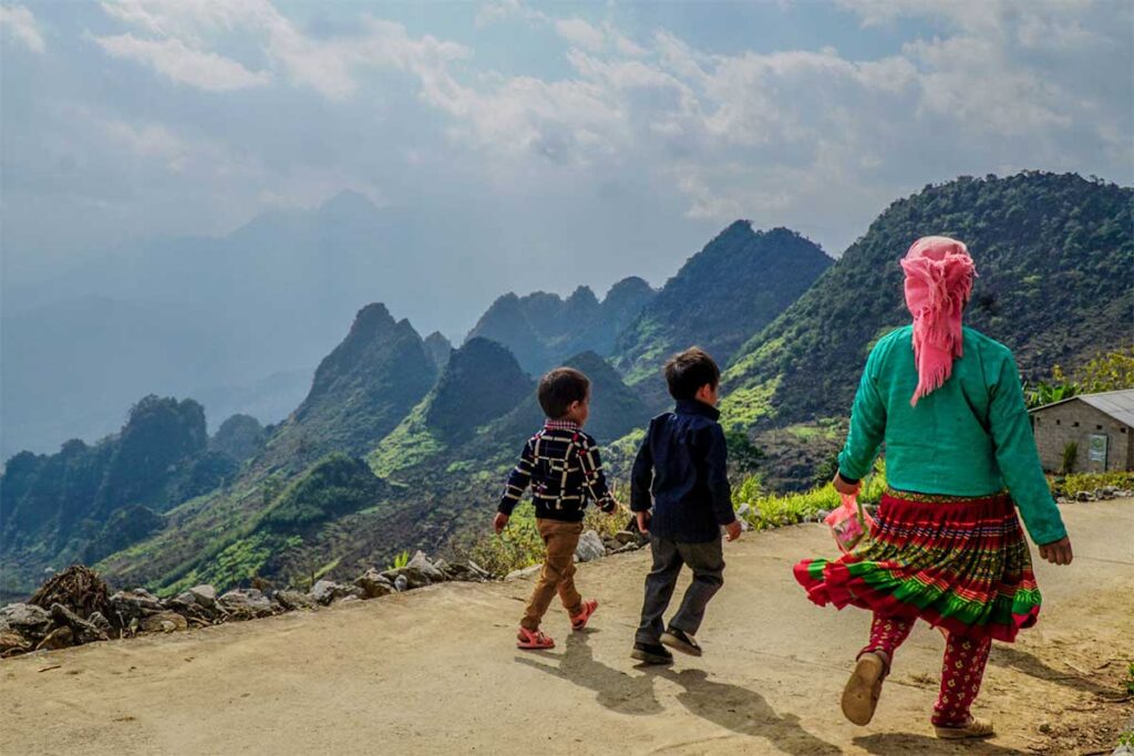 Ethnic minority woman and children walking along a mountain road on the Ha Giang Loop, with dramatic karst landscapes in the background
